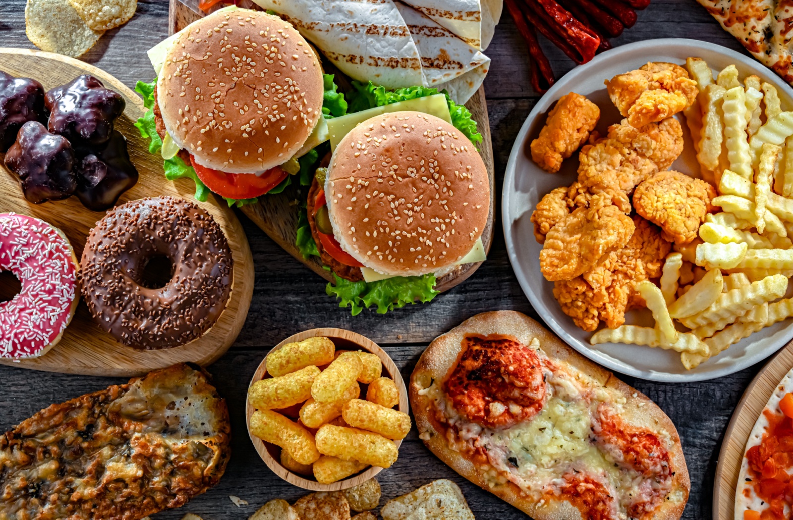 Unhealthy fast foods, including burgers, fries, pizza, and donuts on a wooden table.